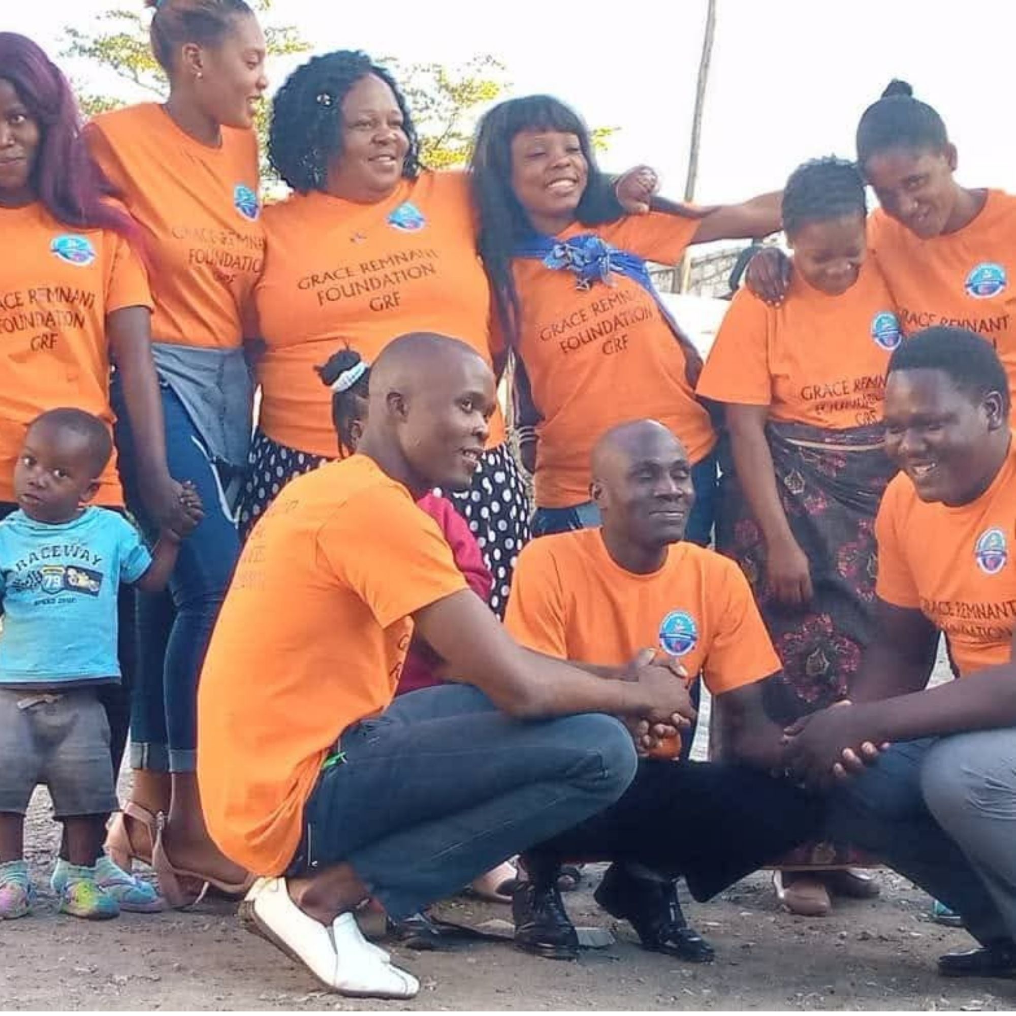 Group of volunteers in orange “Grace Remnant Foundation” t-shirts posing outdoors, with a child in a blue shirt standing on the left.